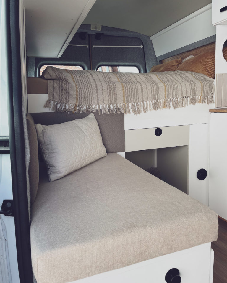 Blick auf die Sitzbank und das Bett mit Kissen und Decken des Kastenwagen Wohnmobils