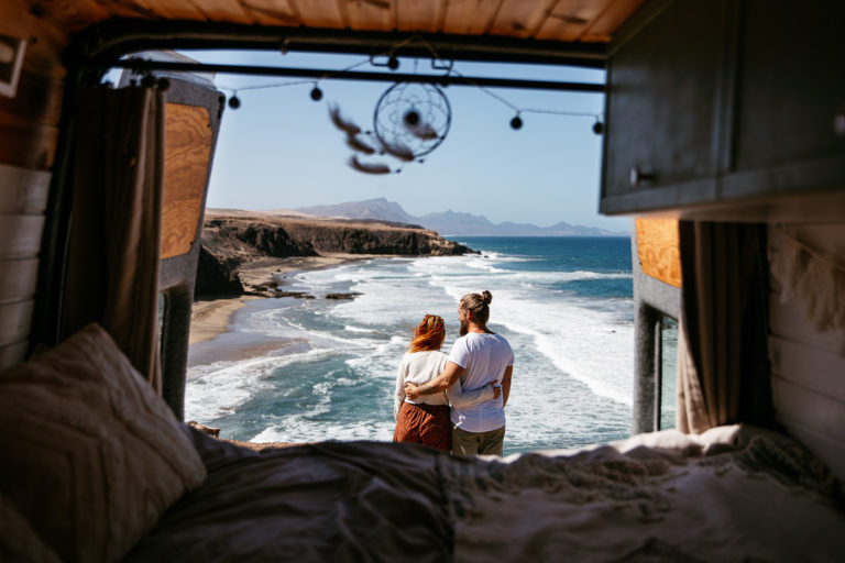 Blick durch die Hecktüren des selbstausgebauten Vans auf die Küste, wo sich ein Paar umarmt